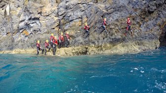 Eine Gruppe von Abenteurern in Neoprenanzügen klettert an einer steilen Felswand entlang, umgeben von klarem, blauem Wasser.