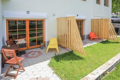 Ein sonniger Außenbereich mit Holzstühlen und bunten Sitzgelegenheiten, umgeben von grünem Gras und einem gepflegten Garten.