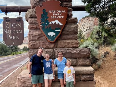 Eine Familie steht lächelnd vor dem Eingang zum Zion Nationalpark, umgeben von beeindruckenden Felsen und Bäumen.