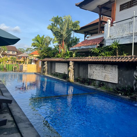 Ein glitzernder Pool spiegelt den blauen Himmel wider, umgeben von üppigem Grün und balinesischer Architektur.