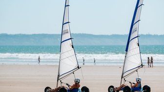 Zwei Kinder fahren in Strandbuggys mit Segeln über den weichen Sandstrand, während das Meer im Hintergrund glitzert.