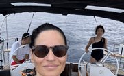 Eine Frau mit Sonnenbrille lächelt in die Kamera, während sie auf einem Segelboot in ruhiger See steht.