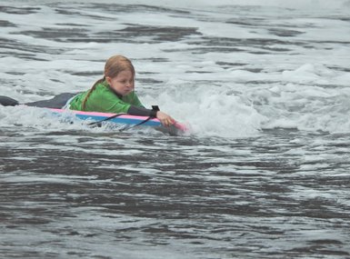 Ein Kind mit langen, blonden Haaren paddelt auf einem Surfbrett durch die Wellen des Ozeans, umgeben von spritzendem Wasser.
