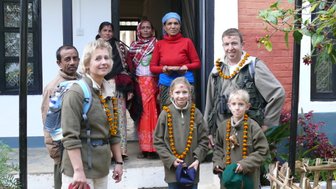 Eine Familie steht vor einem Haus in Nepal, umgeben von Einheimischen, die traditionelle Kleidung tragen und freundlich lächeln.