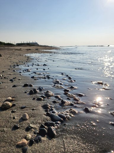 Der Strand ist mit glatten Steinen bedeckt, während sanfte Wellen das Wasser reflektieren und die Sonne strahlt.