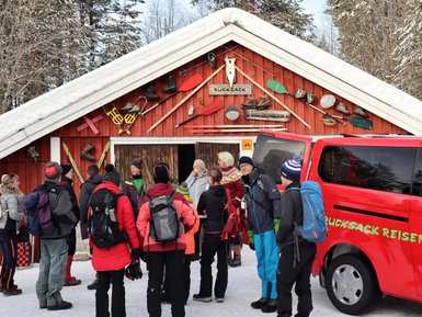 Eine Gruppe von Reisenden steht vor einer roten Hütte, umgeben von schneebedeckten Bäumen, während sie sich unterhalten.