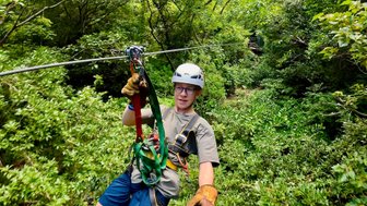 Ein junger Erwachsener, der an einem Zipline-Hochseil durch üppiges, grünes Blattwerk schwingt.