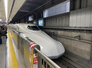 Ein moderner Shinkansen-Zug steht an einem Bahnhof, bereit zur Abfahrt, während Passagiere in der Nähe warten.