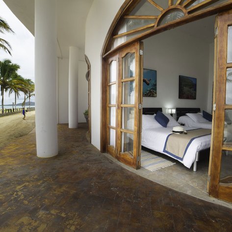 Ein Blick auf ein helles Schlafzimmer mit Holztüren, das auf einen Strand mit Palmen und einem Spaziergänger hinausgeht.