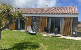 Ein modernes, steinernes Ferienhaus mit Holzjalousien, umgeben von grünem Rasen und einer Palme im Vordergrund.