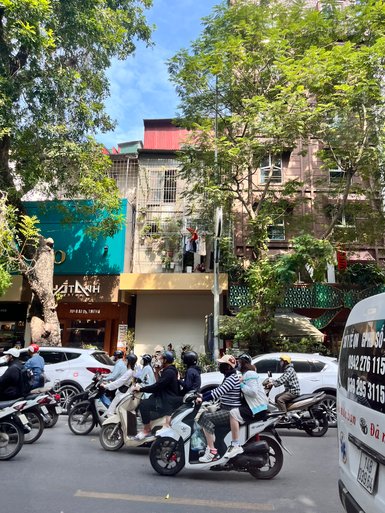 Eine belebte Straße in Hanoi, auf der viele Motorräder und Autos fahren, umgeben von grünen Bäumen und bunten Gebäuden.