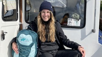 Eine junge Frau mit langen, lockigen Haaren sitzt entspannt auf einem Boot, trägt eine Mütze und eine schwarze Jacke.