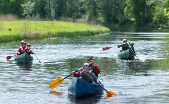 Fünf Personen paddeln fröhlich in Kanus auf einem ruhigen Gewässer, umgeben von üppigem Grün und strahlendem Sonnenschein.