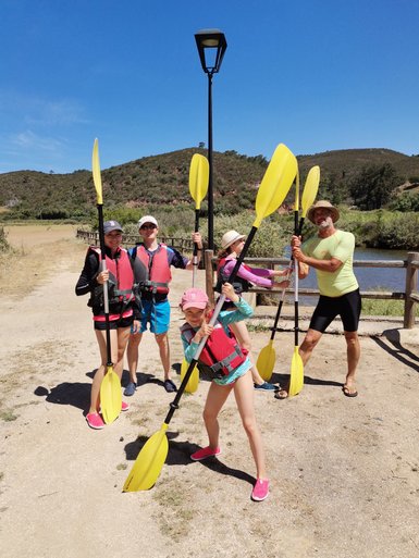 Eine Gruppe von fünf Personen steht am Ufer, bereit zum Kajakfahren, mit Paddeln in der Hand und fröhlichen Gesichtern.