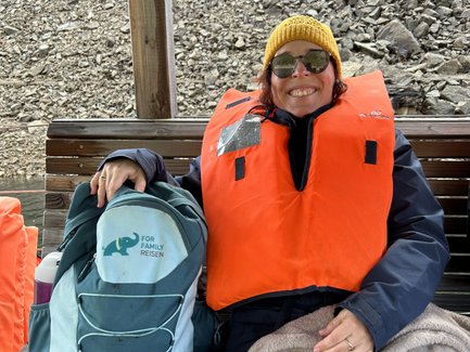 Eine lächelnde Frau in einer orangefarbenen Schwimmweste sitzt auf einer Bank, umgeben von Felsen und Wasser.