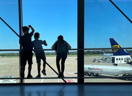 Drei Kinder stehen am Fenster eines Flughafens und beobachten das Geschehen auf dem Rollfeld bei strahlend blauem Himmel.