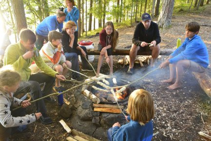 Eine Gruppe von Kindern und Erwachsenen sitzt um ein Lagerfeuer und röstet Teig auf Stöcken in einer Waldumgebung.
