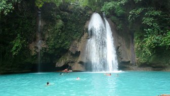 Ein atemberaubender Wasserfall stürzt in ein türkisfarbenes Becken, umgeben von üppigem Grün und fröhlichen Schwimmern.