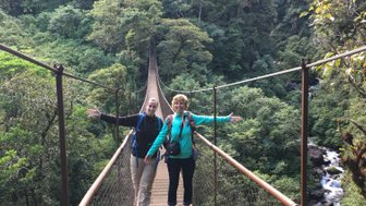 Zwei Wanderer stehen auf einer Hängebrücke, umgeben von üppigem Grün und einem Wasserfall im Hintergrund.