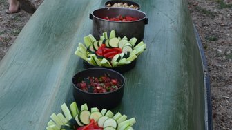 Ein Kanu liegt am Ufer, während frische Salate und Snacks in Schalen auf dem Boot angerichtet sind.