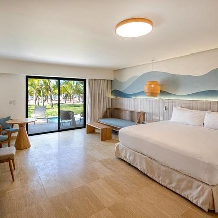 Ein modernes Hotelzimmer mit einem großen Bett, einem Tisch und einem Blick auf die Palmen durch die Fenster.