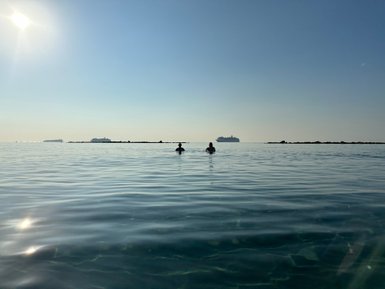 Zwei Personen stehen im ruhigen Wasser, umgeben von einem klaren Himmel und sanften Wellen, während Schiffe in der Ferne sichtbar sind.