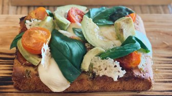 Ein frisch zubereiteter Sandwich mit Tomaten, Avocado und Spinat auf einem Holzbrett, serviert mit einer grünen Sauce.