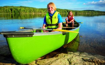 Zwei Kinder sitzen in einem grünen Kanu auf einem ruhigen See, umgeben von üppigem Wald und klarem Himmel.
