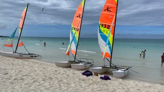 Drei bunte Katamarane stehen am Strand, während Menschen im klaren Wasser planschen und die Wolken am Himmel ziehen.