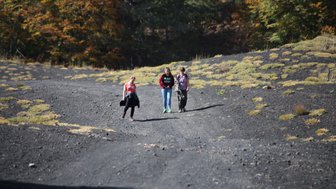 Drei Wanderer gehen auf einem schottrigen Weg, umgeben von herbstlichen Bäumen und grünem Gras.
