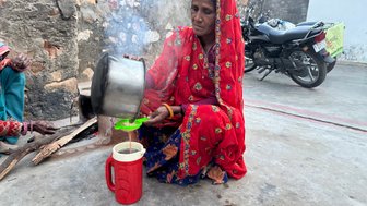 Eine Frau in einem roten Kleid gießt Tee in einen Behälter, während Rauch von einem Feuer aufsteigt.