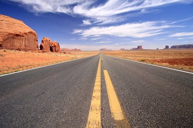 Eine endlose Straße erstreckt sich durch die weite, rote Wüstenlandschaft unter einem strahlend blauen Himmel.
