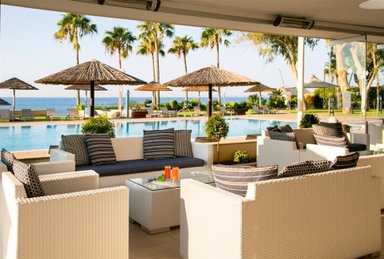 Ein eleganter Außenbereich mit weißen Sofas und einem Glastisch, umgeben von Palmen und einem Pool mit Meerblick.