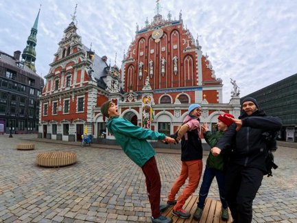 Vier Personen stehen auf einer Holzplattform und posieren fröhlich vor einem historischen Gebäude in Riga.