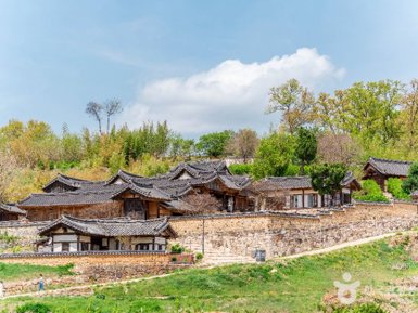 Traditionelle koreanische Häuser mit Ziegeldächern stehen in einer malerischen Landschaft, umgeben von Bäumen und sanften Hügeln.