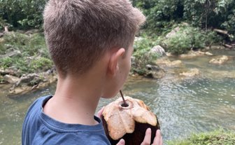 Ein Junge steht am Ufer eines klaren Flusses und hält eine frische Kokosnuss in der Hand, umgeben von üppigem Grün.