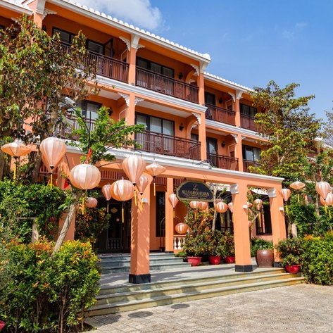 Die Fassade eines Hotels mit orangefarbenem Anstrich, umgeben von üppigem Grün und dekorativen Laternen, strahlt einladende Atmosphäre aus.
