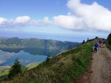 Eine Gruppe von Wanderern geht auf einem schmalen Pfad entlang einer grünen Berglandschaft mit Blick auf einen ruhigen See.