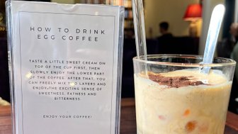 Ein Glas Eierkaffee mit einer Schicht süßer Sahne und einer dunklen Kaffeeschicht, daneben eine Anleitung zum Trinken.