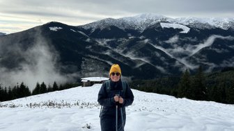 Eine Person steht im Schnee auf einem Berggipfel, umgeben von schneebedeckten Bergen und bewölktem Himmel.
