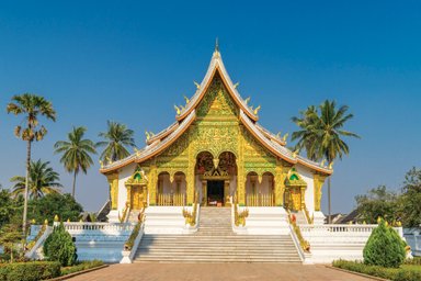 Ein prächtiger Tempel mit goldenen Verzierungen und einer beeindruckenden Treppe, umgeben von Palmen und klarem Himmel.