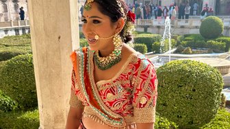 Eine Frau in traditioneller indischer Kleidung lächelt, während sie in einem prächtigen Garten steht, umgeben von historischen Gebäuden.