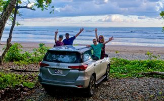 Eine Gruppe von vier Personen steht fröhlich neben einem silbernen Auto am Strand von Costa Rica, umgeben von üppigem Grün.