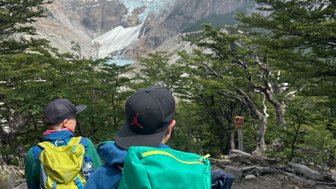 Zwei Wanderer mit Rucksäcken stehen auf einem Pfad, umgeben von Bäumen und Blick auf schneebedeckte Berge im Hintergrund.