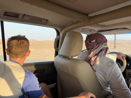 Ein Mann mit einem traditionellen Kopftuch fährt ein Auto durch eine trockene Wüstenlandschaft, während ein junger Mann zuschaut.