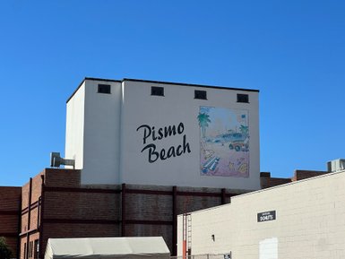 Ein großes Wandgemälde mit der Aufschrift 'Pismo Beach' und einem bunten Strandmotiv ist an einem Gebäude zu sehen.