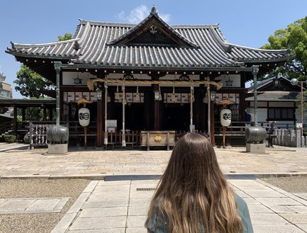 Eine Frau mit langen Haaren steht vor einem traditionellen japanischen Schrein, umgeben von grünen Bäumen und klarem Himmel.