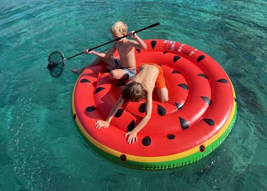 Zwei Kinder spielen auf einem großen, wassermelonengemusterten Schwimmring in klarem, blauem Wasser.