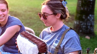 Eine Frau spielt ein traditionelles Musikinstrument im Freien, während eine andere Person lächelt und zuschaut.