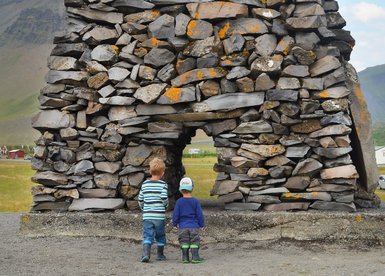 Zwei Kinder stehen vor einem beeindruckenden Steindenkmal, das mit Moos und Flechten bedeckt ist, in einer malerischen Landschaft.